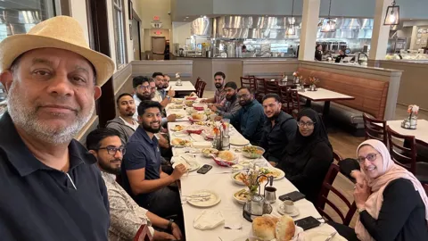 A long dinner table full of guests, with a smiling man in a hat in front.